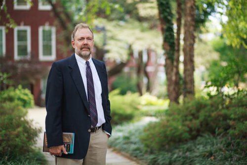 Dr. Nicholas Meriwether is pictured standing on a sidewalk on campus holding a book