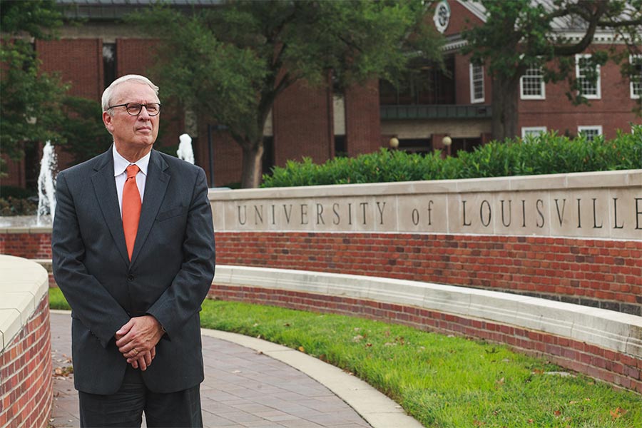 Dr. Allan Josephson stands next to a sign on campus that reads 'University of Louisville.'