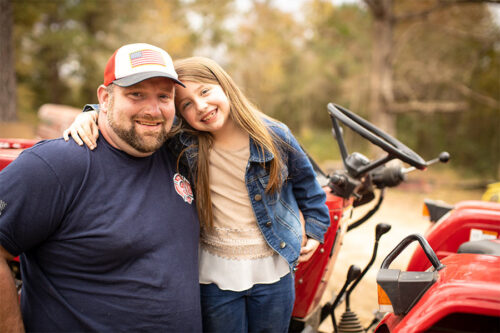 Lydia Booth puts her arm around her dad, Matthew, as the two smile in front of a red tractor.